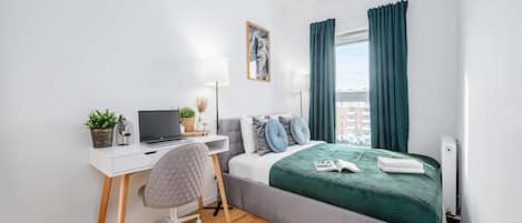 Un dormitorio moderno con una cama, un escritorio con una computadora portátil, una silla y una ventana con cortinas.