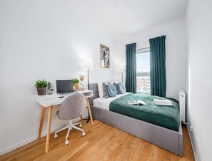 Ein modernes Schlafzimmer mit einem Bett, einem Schreibtisch mit Laptop, einem Stuhl und einem Fenster mit Vorhängen.