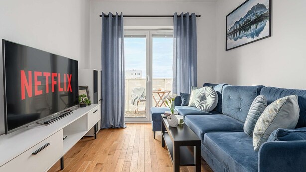 A modern living room with a flat-screen TV displaying 'NETFLIX,' a blue sofa, a coffee table, and a balcony with a view. - Beautiful Seaview Flat in Stagadon Island (Łódź)