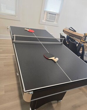 Ein Tischtennis-Tisch mit zwei Schlägern und einem Ball, ein Fenster mit Jalousien und ein Holzboden.