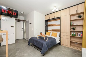 Ein Schlafzimmer mit Bett, Nachttischen, einem Kleiderschrank, einem Bücherregal und einem an der Wand montierten Fernseher mit dem Netflix-Logo.