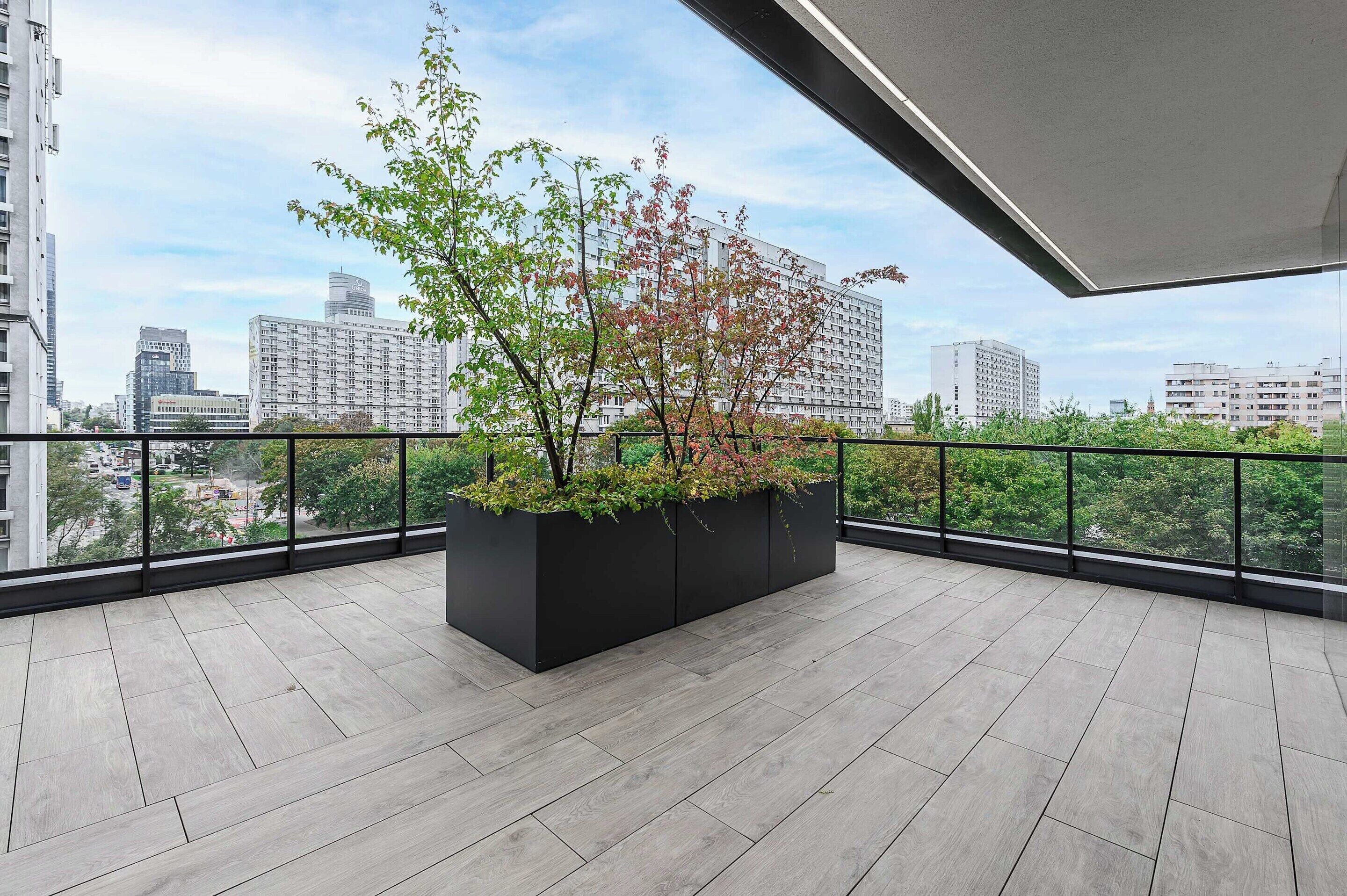 A balcony with a black planter box containing green and red foliage, a tiled floor, and a cityscape view in the background.