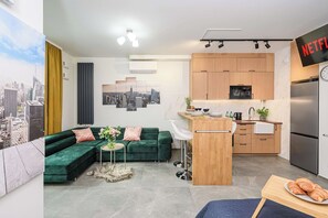 A modern living space with a green sofa, a small round table with a vase, a kitchen area with wooden cabinets, a refrigerator, and a wall-mounted TV displaying the Netflix logo. - Lush Home Near Varso Tower (Warsaw)
