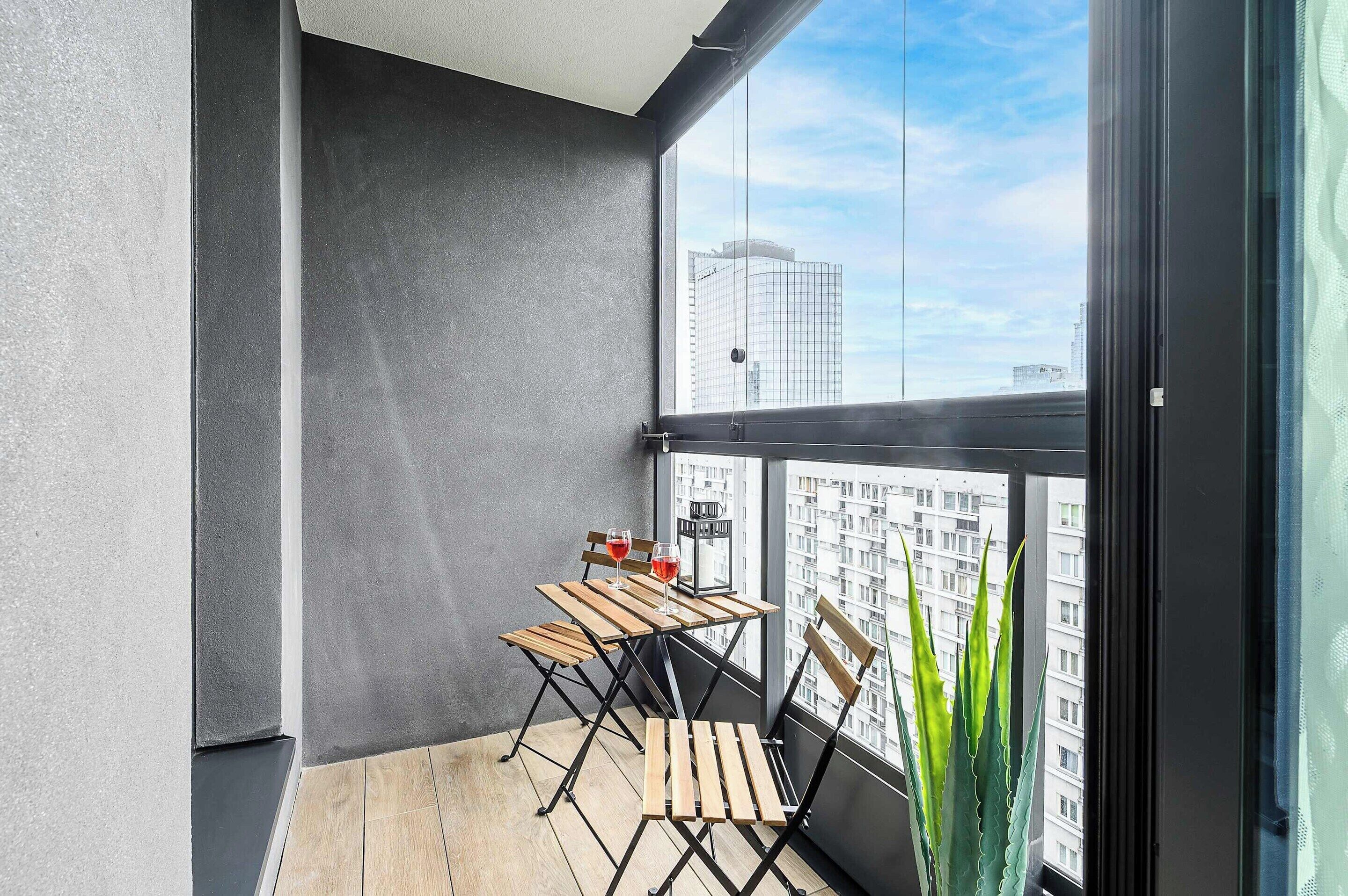 Uma varanda com mesa e cadeiras de madeira, um vaso de planta e vista para a paisagem urbana.
