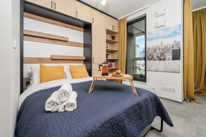 Una habitació d'hotel amb un llit, una taula auxiliar, una biblioteca i una finestra que mostra una vista panoràmica de la ciutat, tot amb una cortina.