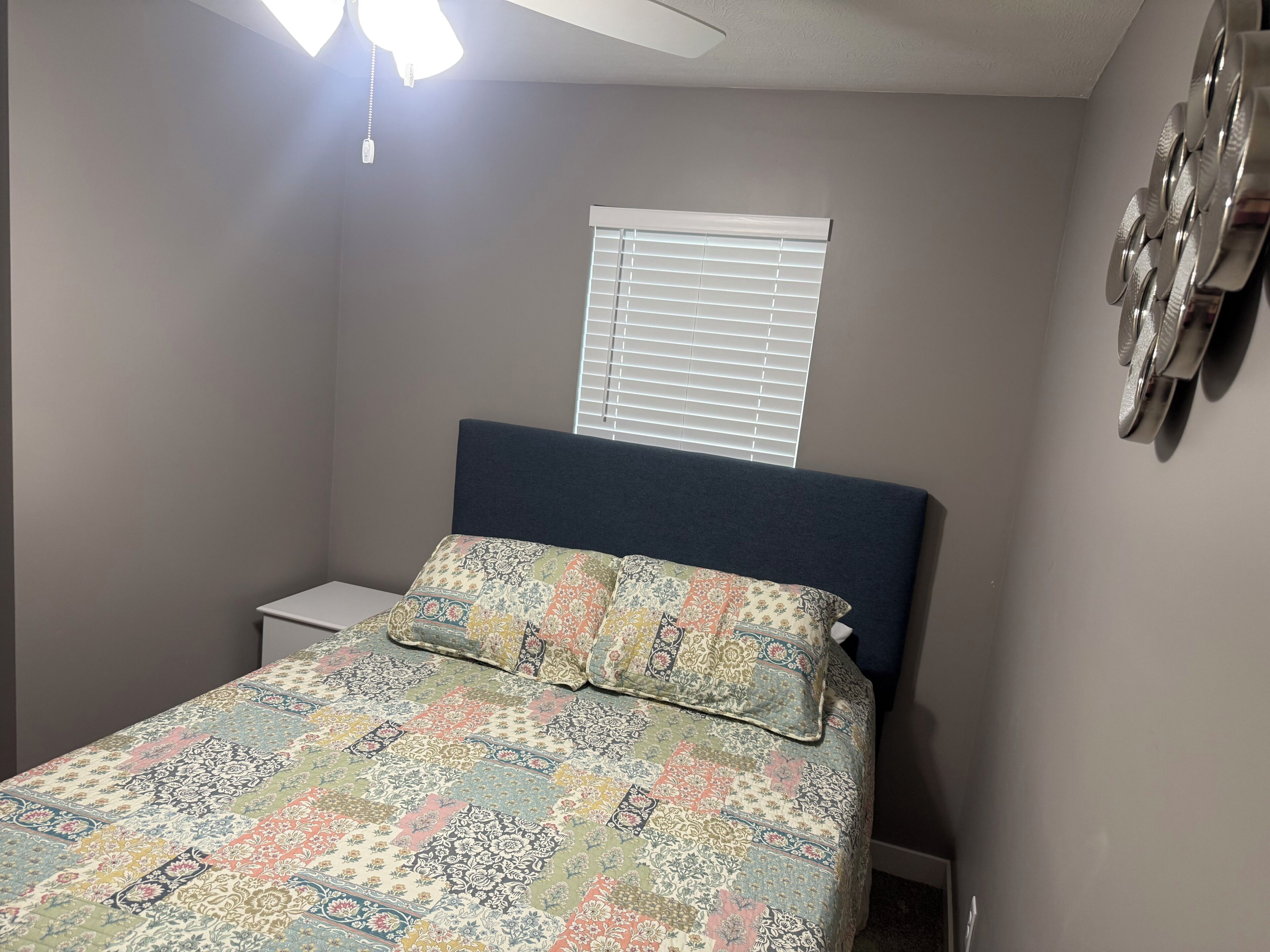 A bedroom with a bed, a nightstand, a window with blinds, and a ceiling fan.