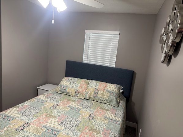 A bedroom with a bed, a nightstand, a window with blinds, and a ceiling fan.