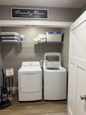 A laundry room with two white front-loading washing machines, a shelf with laundry supplies, and a sign above reading 'Laundry Room Loads of Fun.'