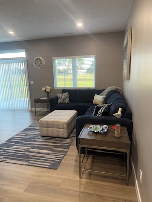 A living room with a dark-colored sofa, a matching ottoman, a coffee table with decorative items, a window with blinds, and a door leading outside.