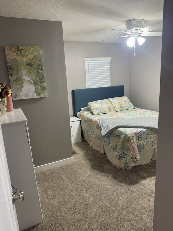 A bedroom with a bed, a nightstand, a dresser, a ceiling fan, and a window with blinds.
