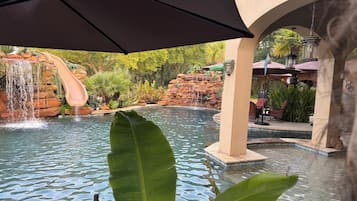 Una piscina con cascada, rodeada de exuberante vegetación y un área de descanso techada.