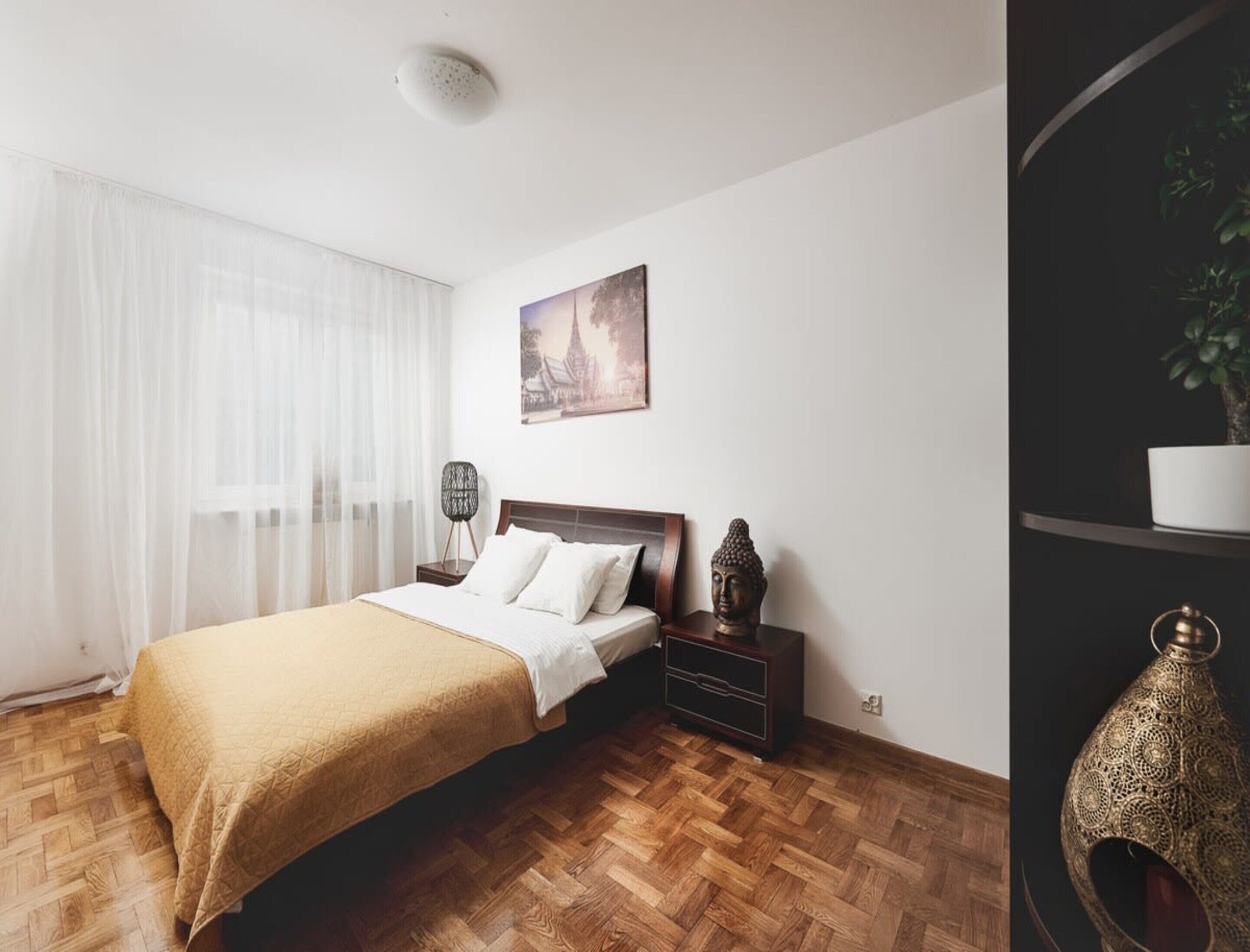Un dormitorio con una cama, una mesita de noche, una lámpara de pared, una estatua de Buda y una planta en maceta.
