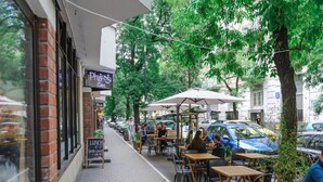 An outdoor café with tables and chairs, a brick building, and parked cars on the street. - Warsaw Home Near Center (Warsaw)