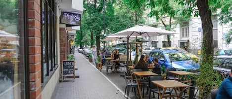 An outdoor café with tables and chairs, a brick building, and parked cars on the street.