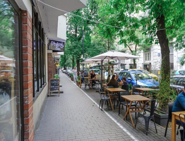 Un café al aire libre con mesas y sillas, un edificio de ladrillo y coches estacionados en la calle.