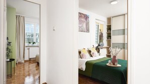 A room with a bed, a radiator, a window with curtains, a shelf with plants, and a door leading to another room. - Warsaw Home Near Center (Warsaw)