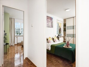Ein Zimmer mit einem Bett, einem Heizkörper, einem Fenster mit Vorhängen, einem Regal mit Pflanzen und einer Tür zu einem anderen Raum.
