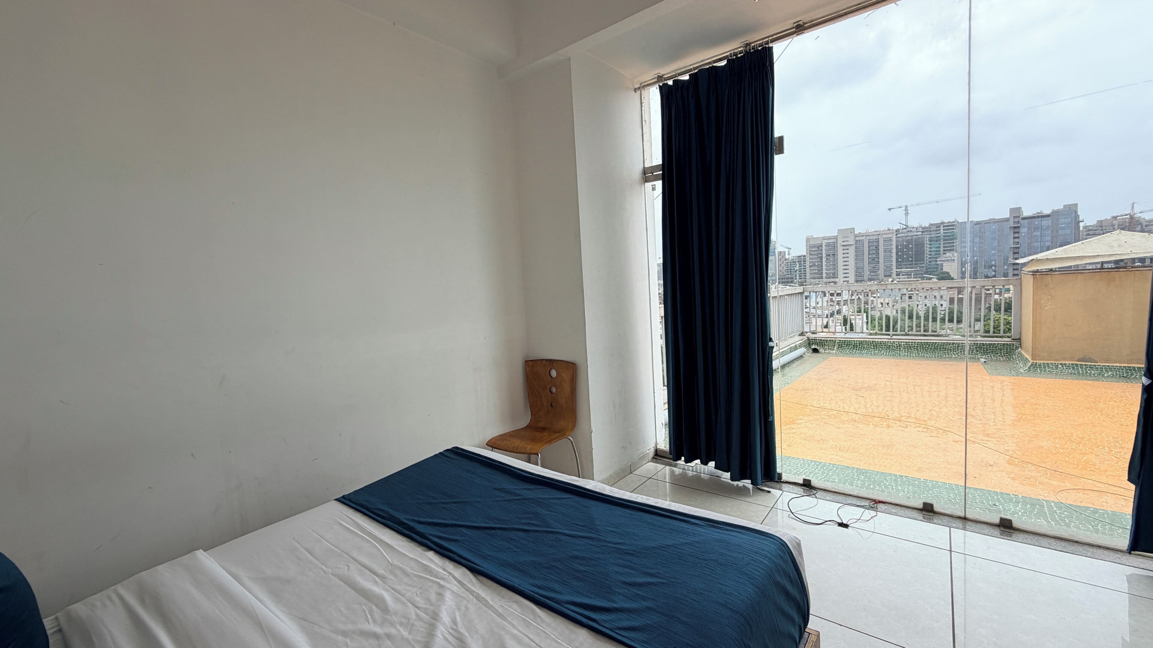 Quarto de hotel com cama, uma cadeira, janela com cortinas e vista para um campo de esportes e edifícios.