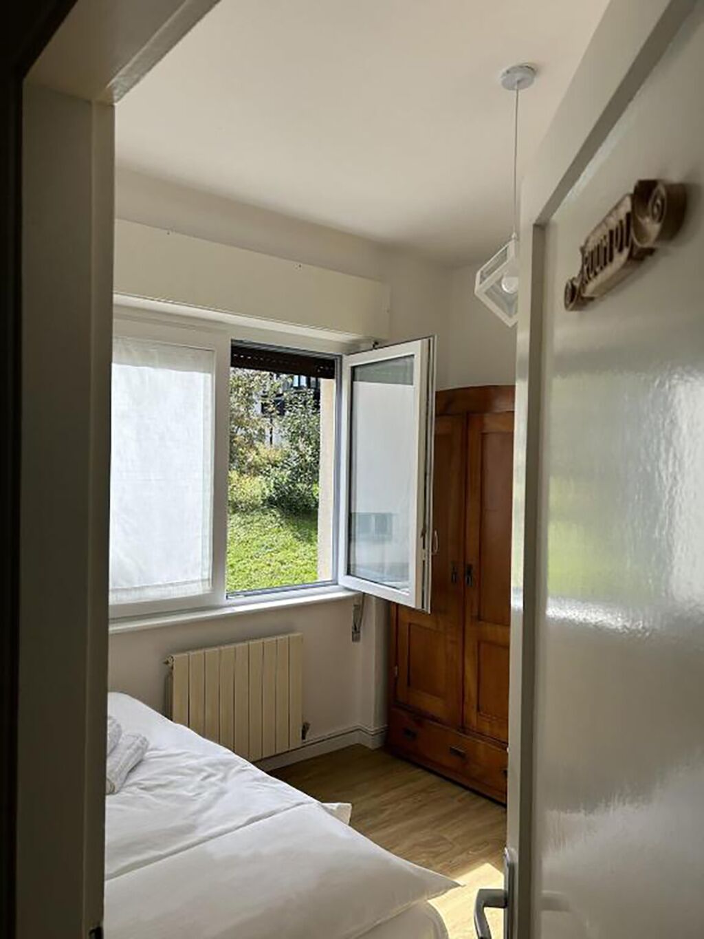 Ein Schlafzimmer mit Bett, Heizkörper, Kleiderschrank und Fenster mit Blick auf Grünflächen.