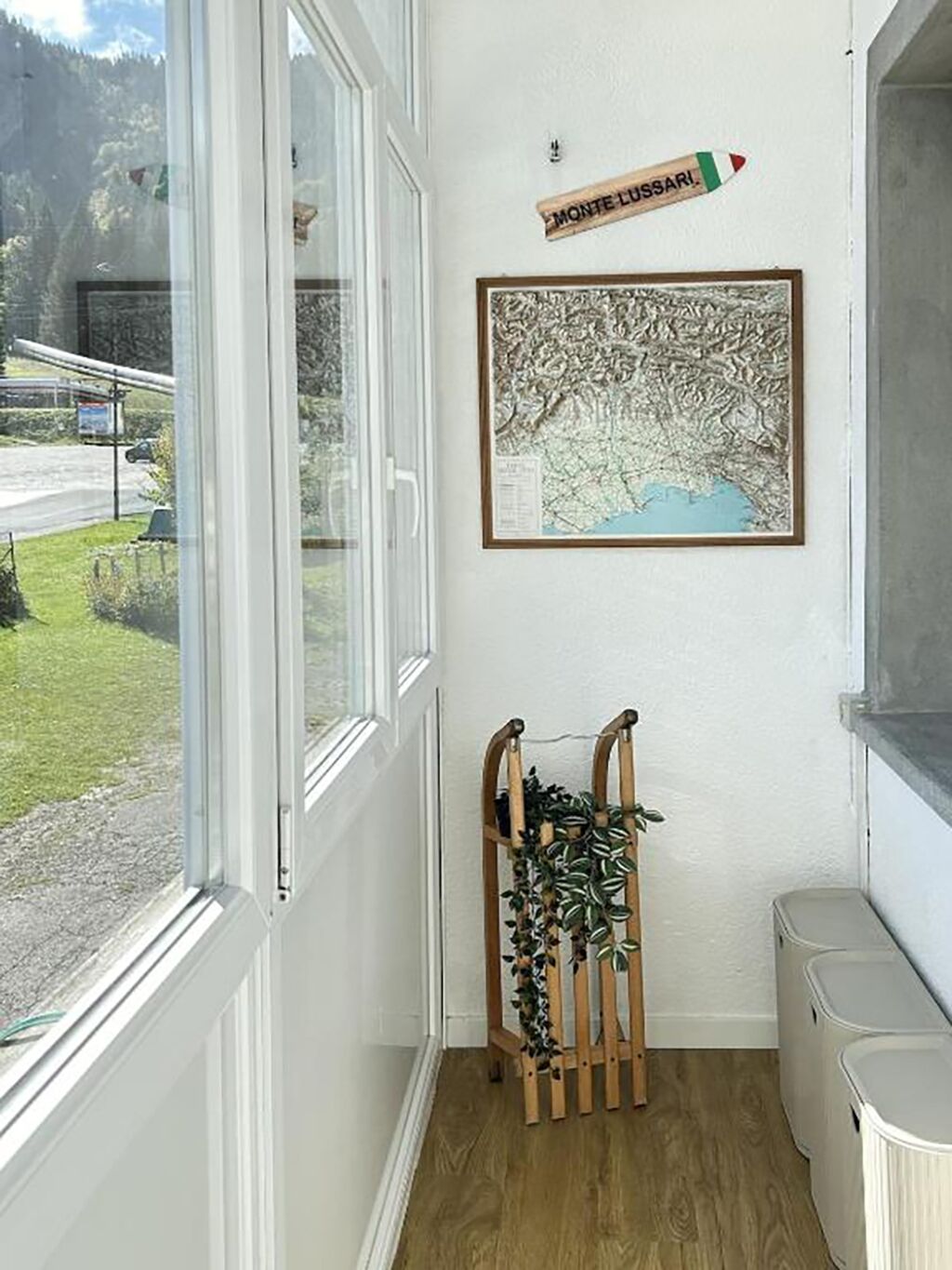 Un couloir avec une fenêtre, une carte encadrée, un présentoir à plantes en bois et une poubelle.
