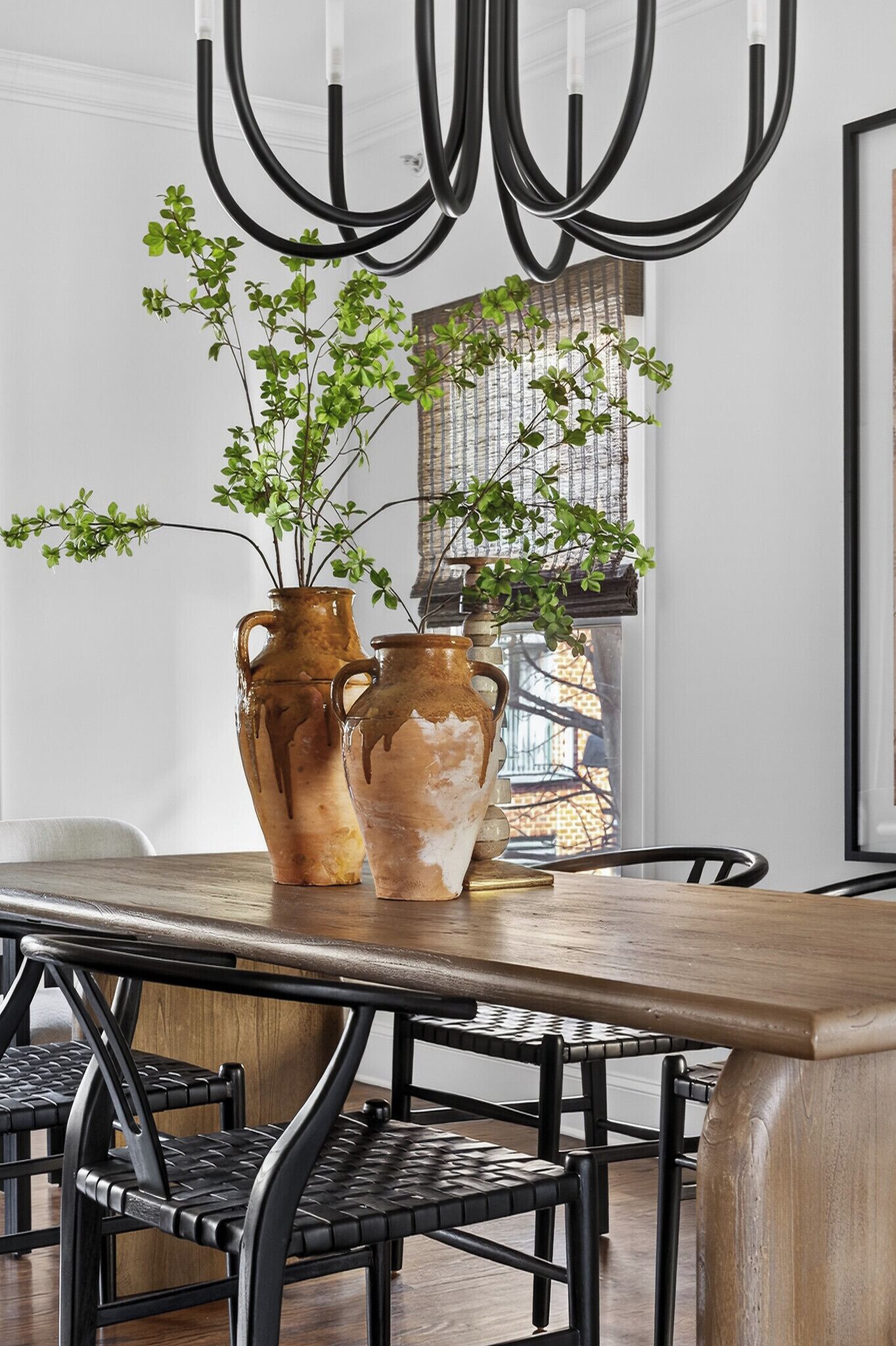 A dining area with a wooden table, two ceramic vases with plants, black chairs, and a modern light fixture.