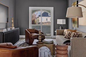 Een woonkamer met een bruine leren bank, een kamerkleed met een patroon, een bijzettafel met boeken, en een lamp.