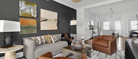 Una sala moderna con un sofá gris, un sillón de cuero marrón y una mesa de centro. Hay pinturas abstractas en la pared y una lámpara de techo tejida.