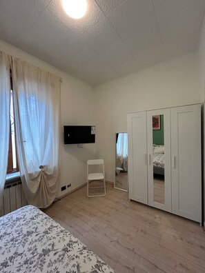 Ein Zimmer mit einem Bett, einem Schrank, einem Sessel, einem Fernseher und einem fenster mit Vorhängen.