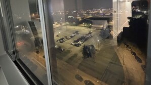 Vista noturna da janela com vista para um estacionamento, vários carros e o horizonte da cidade ao longe.