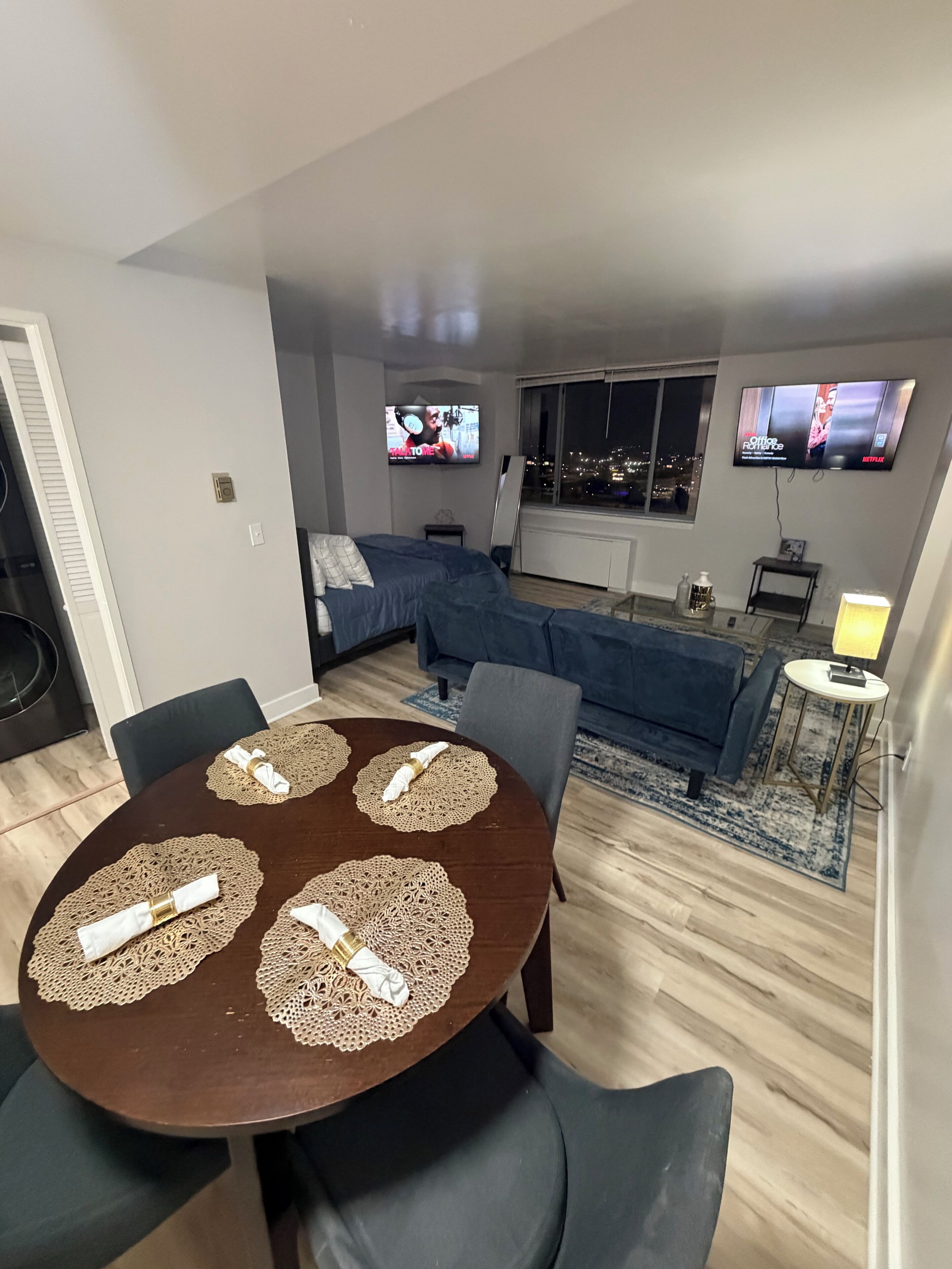 Un salón moderno con una mesa de comedor preparada para cuatro personas, un sofá, dos televisores de pantalla plana montados en la pared y vistas a la ciudad desde la ventana.