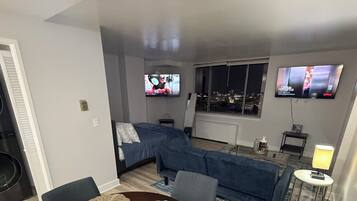 Un salón moderno con una mesa de comedor preparada para cuatro personas, un sofá, dos televisores de pantalla plana montados en la pared y vistas a la ciudad desde la ventana.