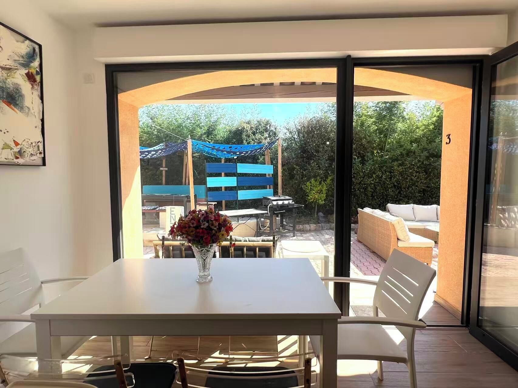 A room with a white table and chairs, a vase with flowers, and a view of an outdoor patio with a barbecue and seating area.