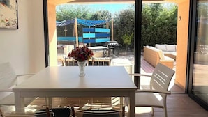 A room with a white table and chairs, a vase with flowers, and a view of an outdoor patio with a barbecue and seating area. - Guesthouse with a pool, 200 meters from the beach, air-conditioned (Roquebrune-sur-Argens)