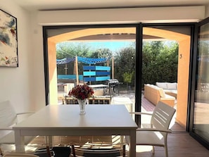 Una habitación con una mesa y sillas blancas, un florero con flores y vista a un patio exterior con una barbacoa y zona de estar.