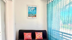 A room with a dark-colored sofa, two patterned cushions, a framed picture on the wall, and a window with blue curtains. - Guesthouse with a pool, 200 meters from the beach, air-conditioned (Roquebrune-sur-Argens)