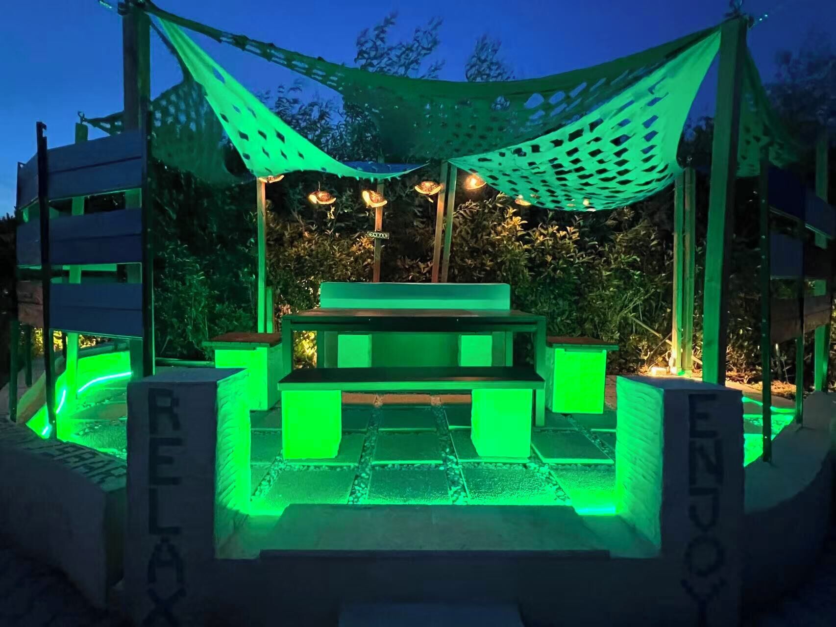 An outdoor seating area with a table and benches under a green canopy, surrounded by greenery.