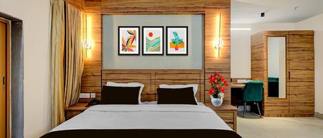 A hotel room with a wooden headboard, a bed with black and white bedding, a desk with a chair, and a mirror. There are three framed artworks on the wall.