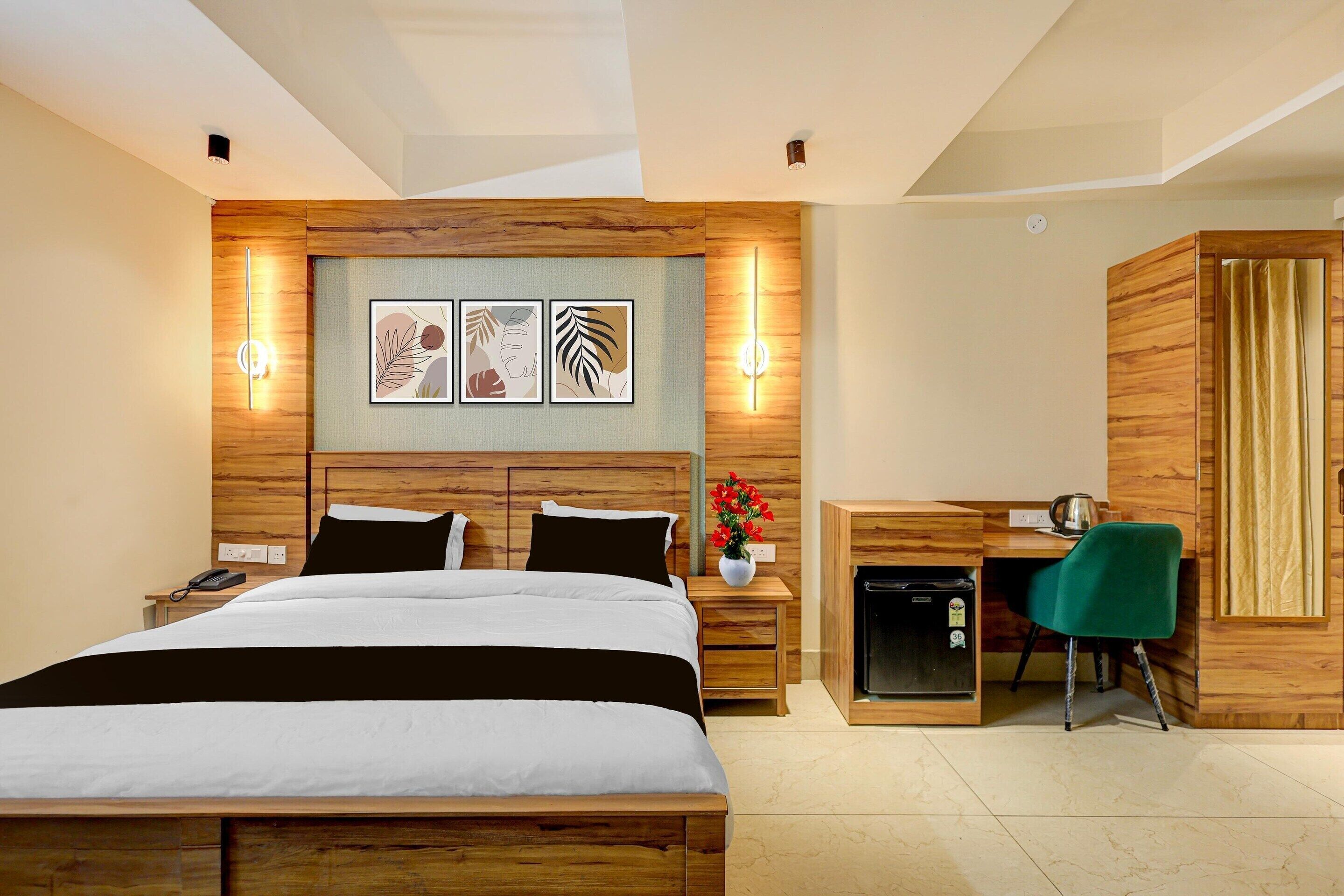 A hotel room with a bed, bedside table, lamp, artwork, desk, chair, and a small refrigerator.