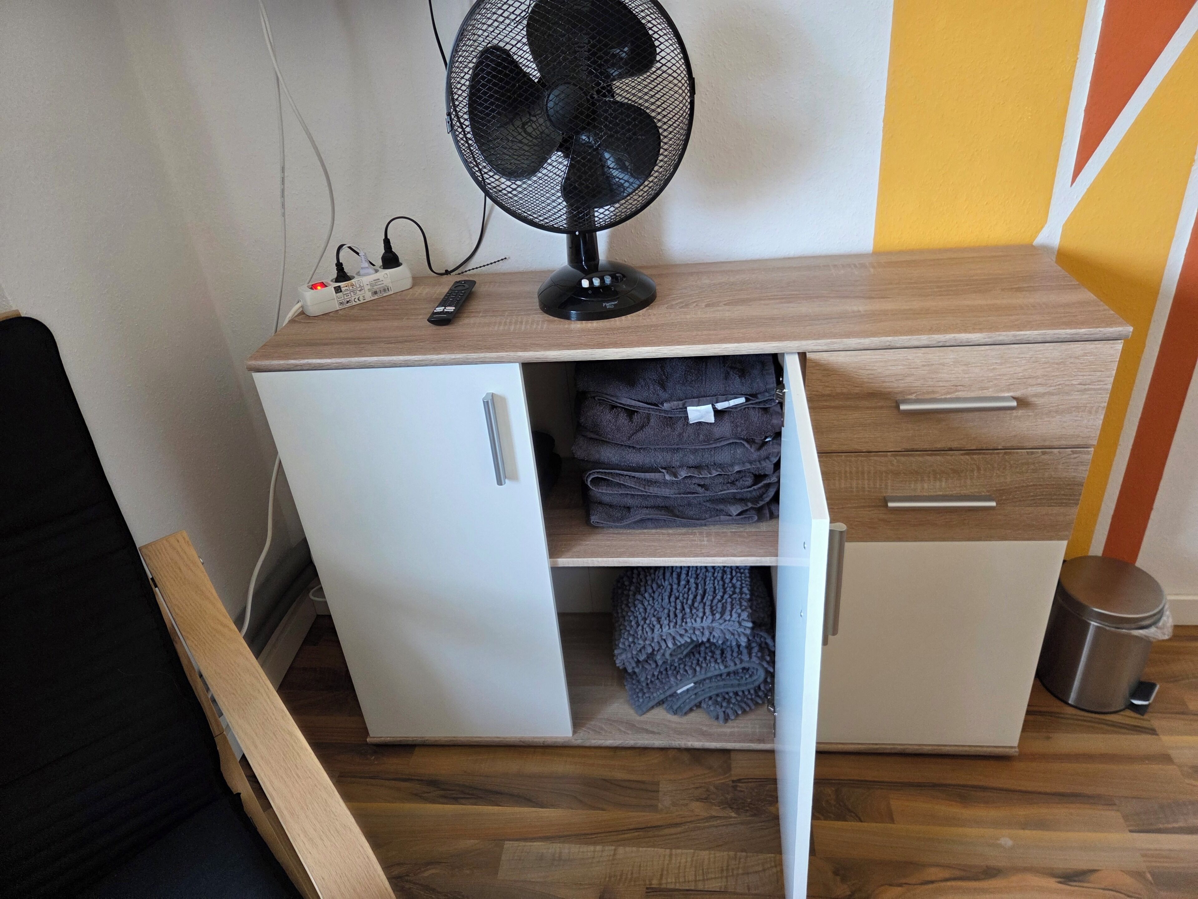 Une pièce de taille modeste, avec un meuble blanc, un ventilateur sur pied et une télécommande posée sur un bureau en bois.
