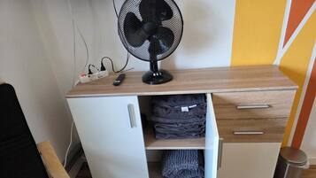 Une pièce de taille modeste, avec un meuble blanc, un ventilateur sur pied et une télécommande posée sur un bureau en bois.