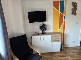 Una habitación con una silla negra, un armario blanco con cajones, un televisor montado, un ventilador de pie y un cubo de basura.