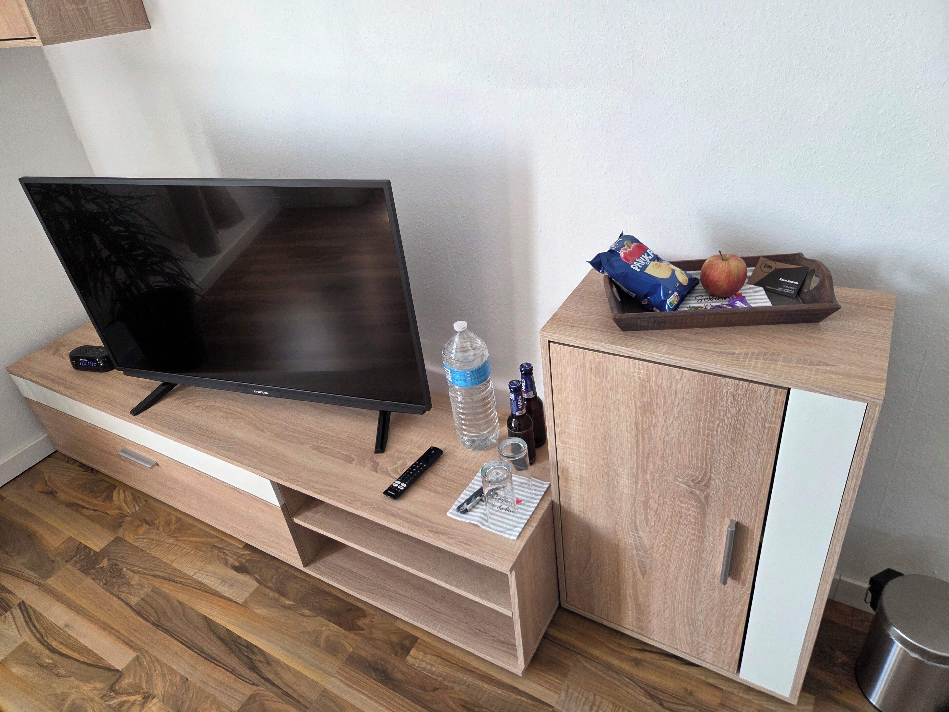 Un meuble en bois pour la télévision, sur lequel est posée une télévision à écran plat, ainsi que des télécommandes, une bouteille d’eau, un verre et un plateau contenant des en-cas et une carte.