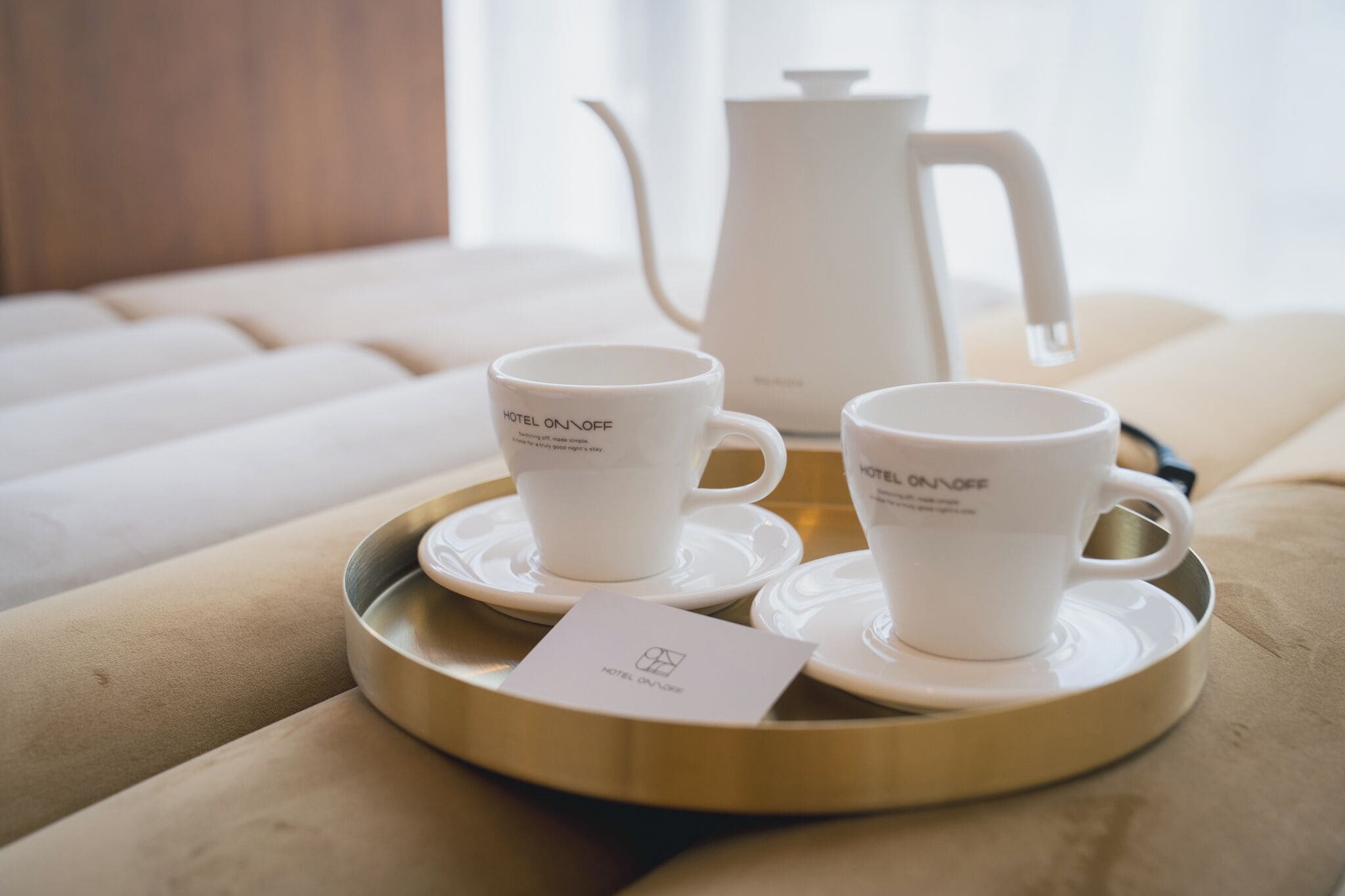 Two white cups with 'HOTEL ON OFF' text on a tray with saucers, a teapot, and a menu card on a beige sofa.