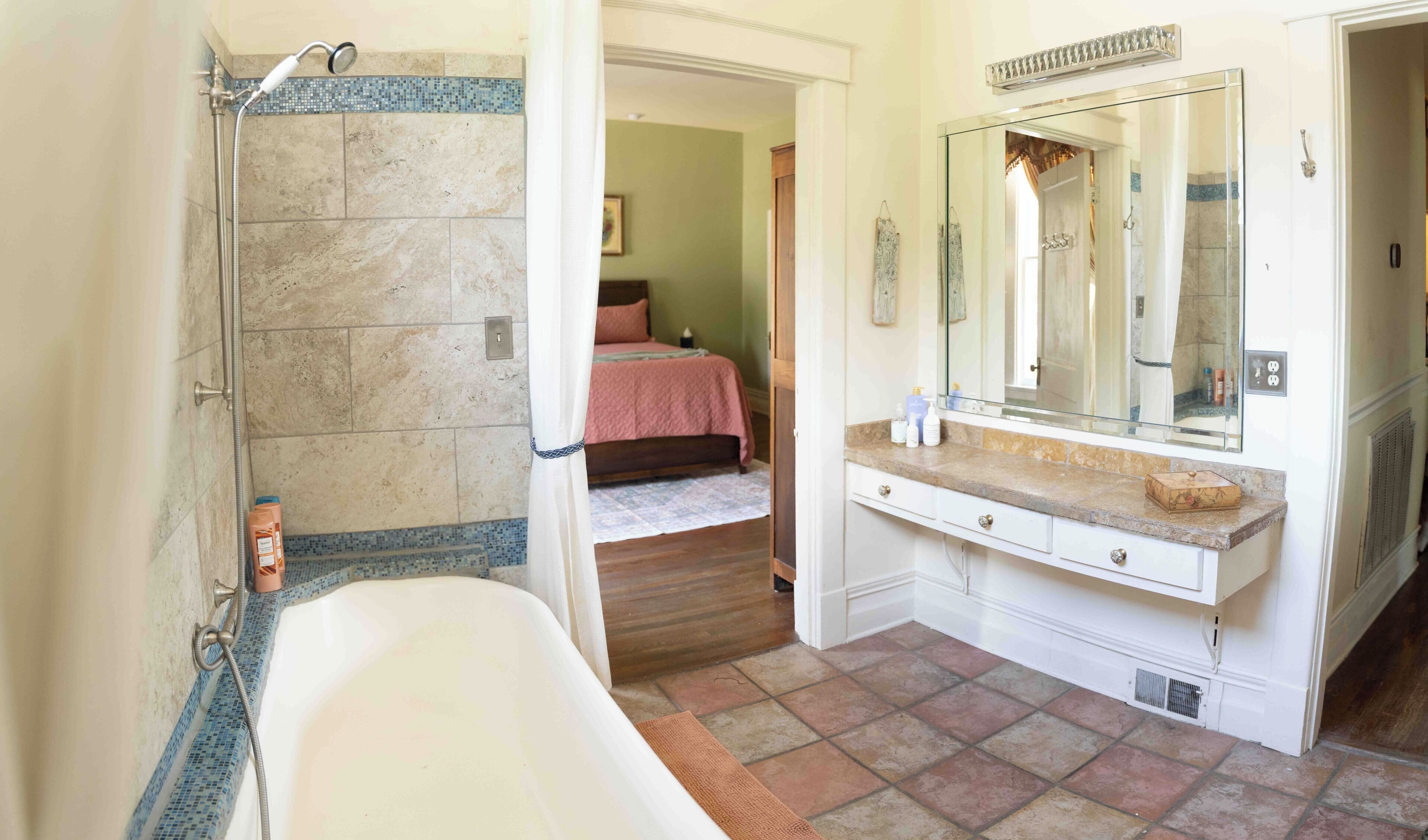 A bathroom with a bathtub, a shower, a vanity with a mirror, and a bed visible through an open door.