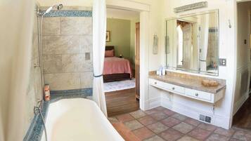 A bathroom with a bathtub, a shower, a vanity with a mirror, and a bed visible through an open door.