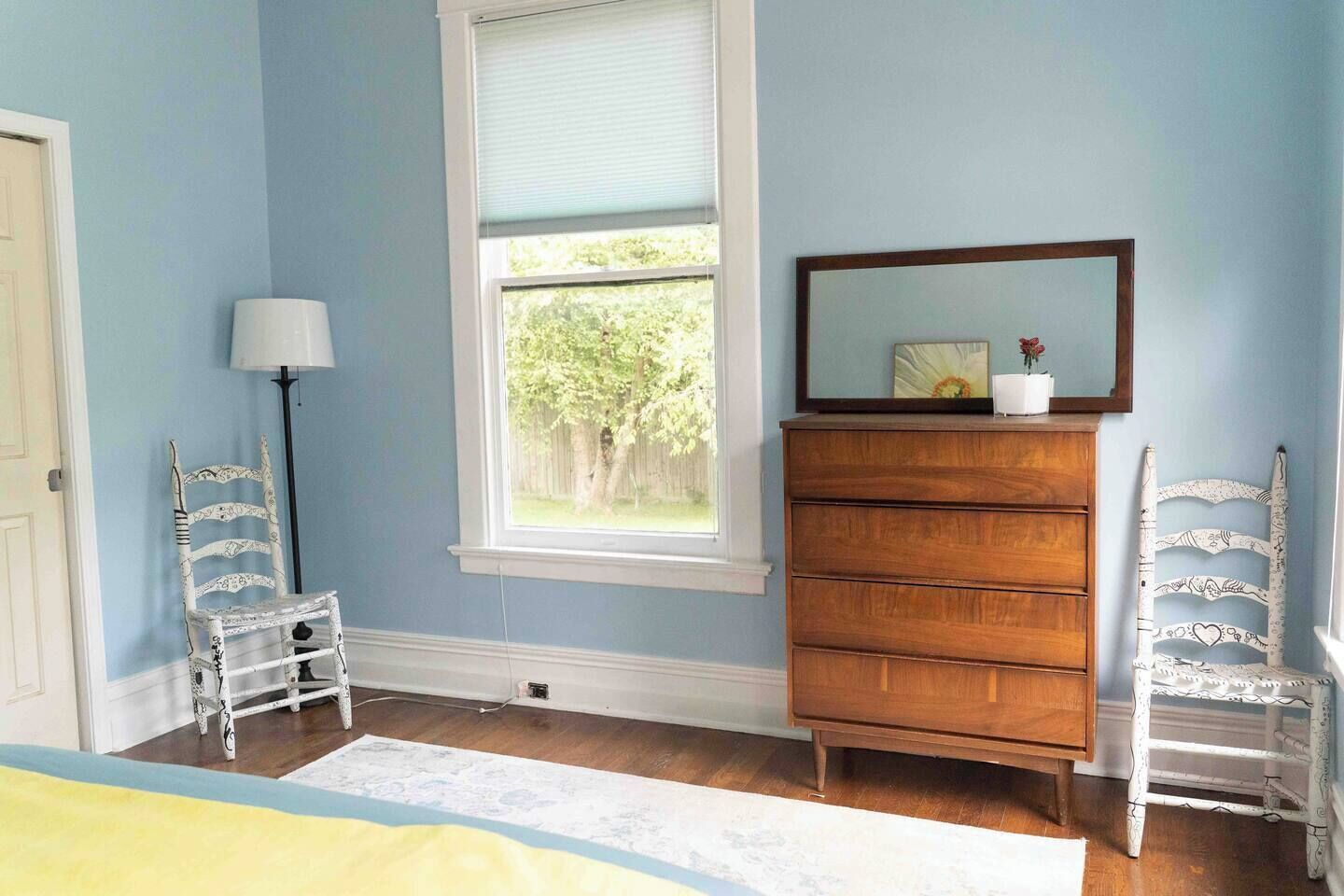 A room with a wooden dresser, a chair, a lamp, and a window with a view of greenery.