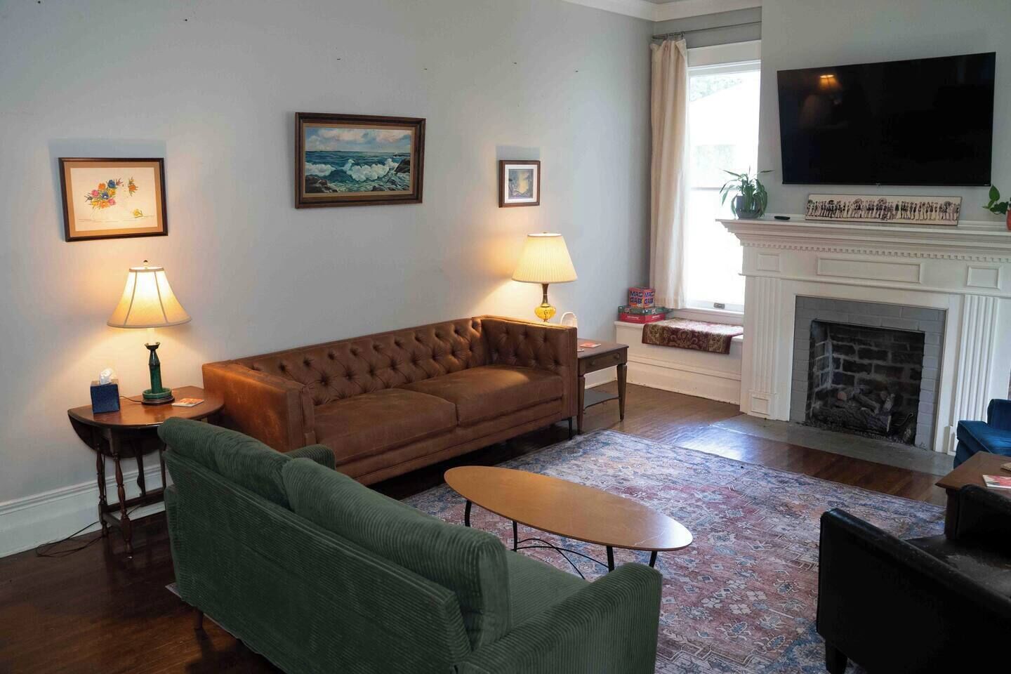 A living room with a brown sofa, green armchair, and a small table. There are two lamps, a fireplace, and a television mounted on the wall.