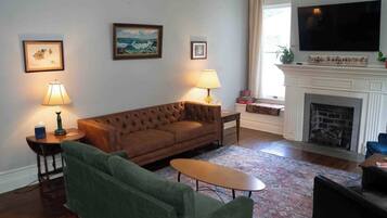 A living room with a brown sofa, green armchair, and a small table. There are two lamps, a fireplace, and a television mounted on the wall.