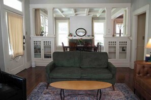 A living room with a green sofa, a wooden coffee table, and a patterned rug. There are white cabinets with glass doors, a dining table with chairs, and a fireplace with a mirror above it.