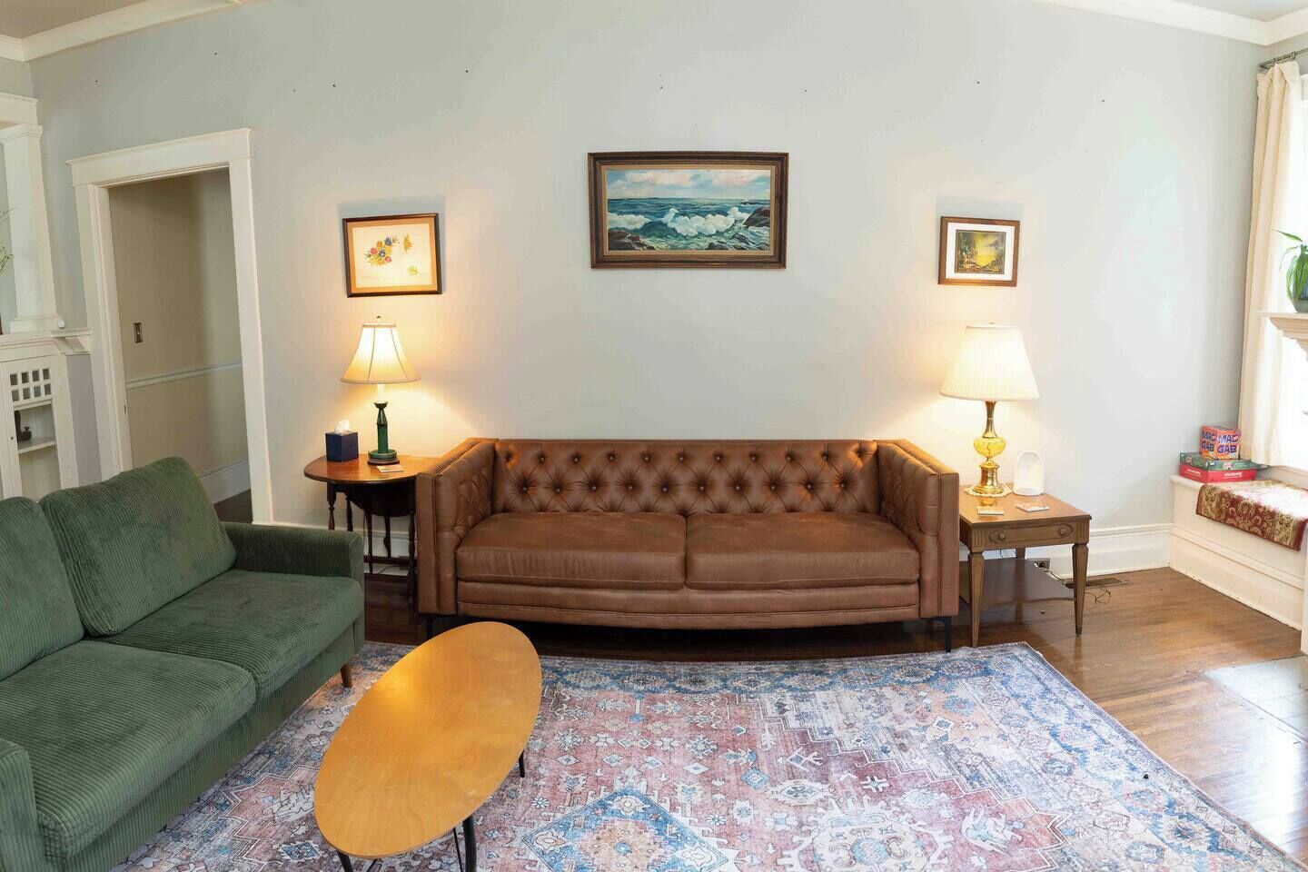 A living room with a brown sofa, a green armchair, a wooden coffee table, and a patterned rug. There are two lamps on side tables and a framed painting on the wall.